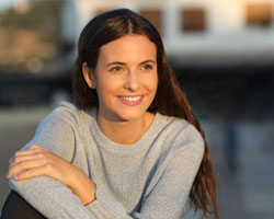 A young female wearing a gray sweater and seated outside while smiling as the sun sets after seeing her Delta Dental dentist in Dallas